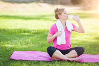 Young fit adult woman outdoors on her yoga mat with towel drinking from her water bottle