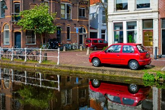 Cars and bicycles on canal embankment in street of Delft with reflection