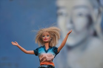 Barbie doll with the gesture of outstretched arms in front of wall with Buddha head