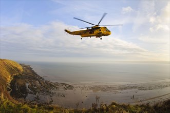 RAF Westland Sea King rescue helicopter with crew members standing in an open doorway in flight over rocky shores and cliffs