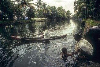 A man kayaking on backwaters and a woman bathing her child the bank of backwater in Kodungallur