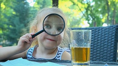 Little girl looks into the lens around. Close-up of blonde girl studying the world around her looking at it through magnifying glass while sitting in street cafe on the park. Odessa