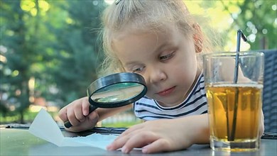 Little girl carefully looks into the lens at the salt. Close-up of blonde girl is studying salt crystals while looking at her through magnifying glass while sitting in street cafe in the park. Odessa