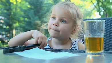 Cute little girl sits thoughtfully at the table with a lens in her hand. Close-up of blonde girl holding a magnifying glass in her hand and thinking about something while sitting in street cafe on the...