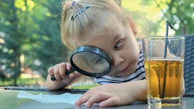 Little girl carefully looks into the lens at the salt. Close-up of blonde girl is studying salt crystals while looking at her through magnifying glass while sitting in street cafe in the park
