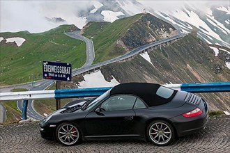 Porsche 911 cabriolet with closed top stands in front of official information sign Edelweissgrat 2460 metres above sea level on pass road from mountain pass above tree line in High Alps