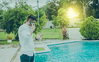 Attractive guy standing talking on the phone near the pool edge. Handsome man calling on the phone by a swimming pool