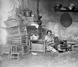 Poor Mexican woman sitting in her kitchen at the charcoal kettle and next to her is a stack of woven baskets