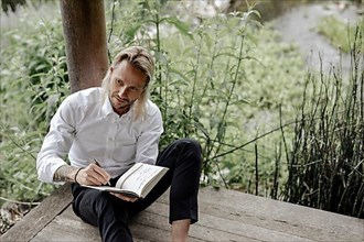 Businessman in white shirt sits on wooden jetty and writes in a book,