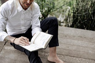 Businessman in white shirt sits on wooden jetty and writes in a book,