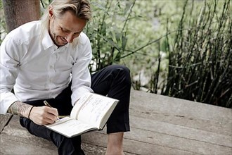 Businessman in white shirt sits on wooden jetty and writes in a book,