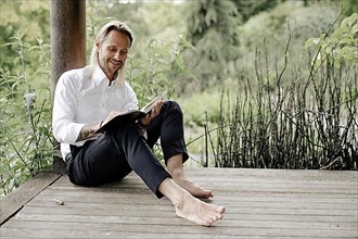 Businessman in white shirt sits on wooden jetty and writes in a book,