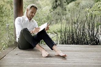 Businessman in white shirt sits on wooden jetty and writes in a book,
