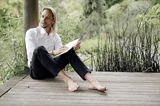 Businessman in white shirt sits on wooden jetty and writes in a book,