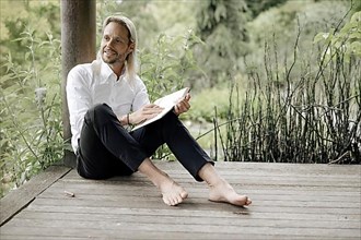 Businessman in white shirt sits on wooden jetty and writes in a book,