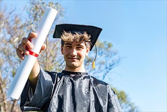 Happy graduated young man wearing a bachelor gown and a black mortarboard and showing his diploma,