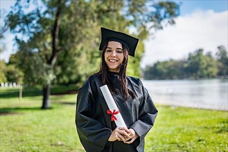 Happy caucasian graduated girl with long brown hair. She is wearing a bachelor gown and a black mortarboard. She is holding a diploma,