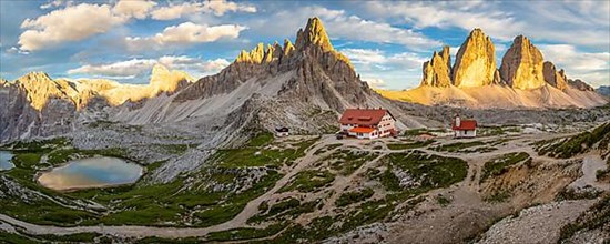 View of mountain hut Dreizinnenhuette and chapel with mountain lake rock massif Paternkofel and mountains three peaks in the sunset, panorama