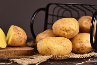 Raw potatoes on wooden cutting board with more potatoes in metal basket in background,