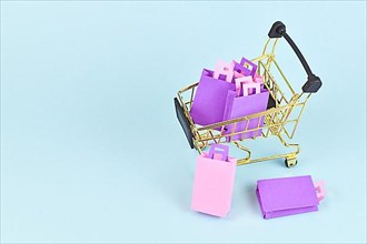 Small golden shopping cart filled with pink and purple paper shopping bags on side of blue background with copy space,