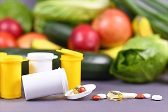 Pills of food nutrition supplements spilling out of bottle in front of fruits and vegetables in background,
