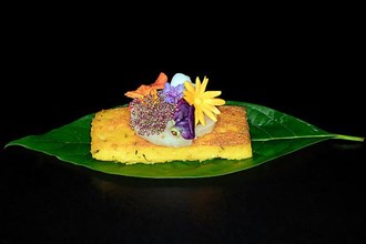 Fried polenta slice with a slice of lotus root and meadow flowers as decoration on an avocado leaf, foddfotography with black background