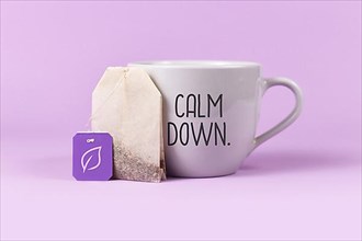 Concept for calming herbal tea with tea bag with leaf on label next to tea cup with text Calm down on violet background,