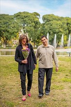 Mature transgender woman walking hand in hand with her boyfriend in a park happy because he gave her flowers,
