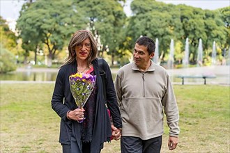 Mature transgender woman walking hand in hand with her boyfriend in a park happy because he gave her flowers,