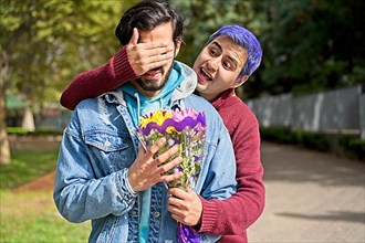 Couple of Latino gay men in a park embracing each other. One surprising the other with a bouquet of flowers as a gift,