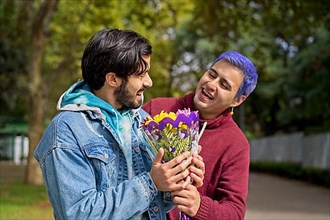 Couple of Latino gay men in a park embracing each other. One surprising the other with a bouquet of flowers as a gift,