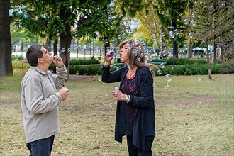 Cheerful mature queer couple blowing bubbles in a park,
