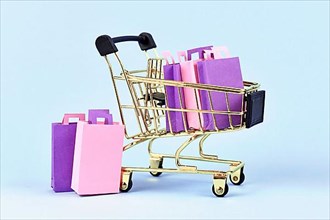 Golden shopping cart filled with pink and purple paper shopping bags on blue background,
