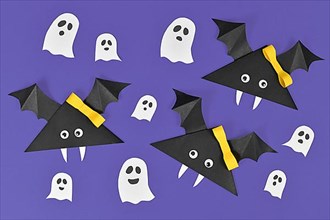 Paper vampire bats with funny googly eyes and ribbons and ghosts on purple Halloween background,