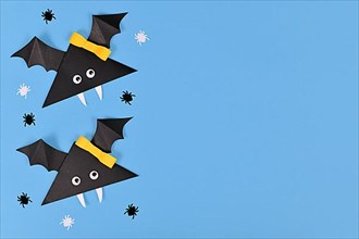 Two cute paper vampire bats with funny googly eyes and ribbon surrounded by small spider confetti on side of blue Halloween background,