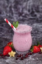 Fruit smoothie shake in drinking glass with striped straw surrounded by ingredients like strawberry and banana pieces and pomegranate seeds on dark background,