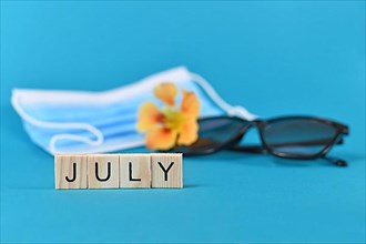 Concept for month July with wooden letters and blurry sunglasses, flower and medical face mask in blue background