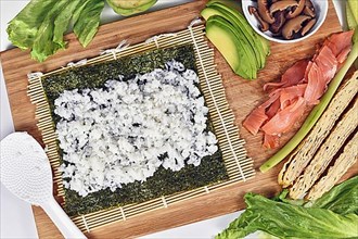 Homemade sushi food preparation with Nori algea sheet with rice on bamboo mat and ingredients like smoked salmon, avocado and egg omelet