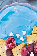 Detail shot of healthy fruit smoothie bowl dyed blue with natural spirulina powder and topped with red raspberry and cranberry and small biscuits,