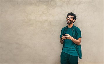 Happy latin man with his cell phone leaning against a wall with copy space, Person smiling phone with space for text. Happy handsome guy leaning against the wall using his cell phone