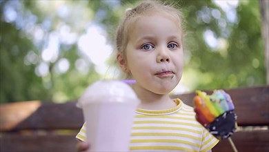 Ittle girl eats a colorful gingerbread and holds a milkshake in her hand. Close-up of cute child girl sitting on park bench and eating cookies with a milkshake and looks into the camera lens. Odessa, ...