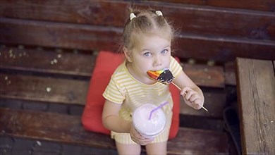 Ittle girl eats a colorful gingerbread and holds a milkshake in her hand. Close-up of cute child girl sitting on park bench and eating cookies with a milkshake and looks into the camera lens. Top view...