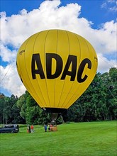 Hot air balloon with inscription ADAC stands with basket on landing field meadow, next to it passengers wait for boarding