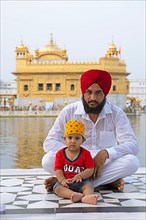 Indian Sikh in white suit and barefoot with his son sitting at the edge of the water basin of the Amrit Sagar or Sacred Lake, at the Hari Mandir or Golden Temple