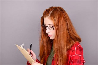 Woman with red hair and glasses looking at clipboard with pen in hand in front of gray wall