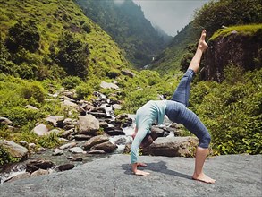 Vintage retro effect hipster style image of woman doing yoga asana eka pada urdva dhanurasana Upward Bow Pose outdoors at waterfall in Himalayas
