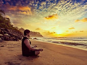 Vintage retro effect hipster style image of sporty fit man doing yoga meditating in padmasana lotus pose on tropical beach on sunset