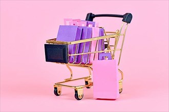Golden shopping cart filled with pink and purple paper shopping bags on pink background