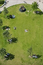 Aerial view over people sunbathing and sitting in the shade of trees in small green municipal park in summer