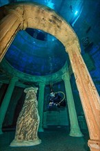 Diver dives through stylised ruin of antique temple round temple in 20 metres water depth in indoor diving tower